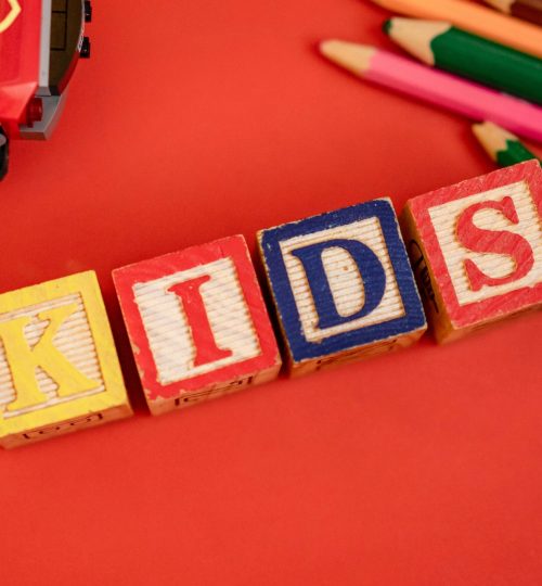 Creative layout of wooden blocks spelling 'KIDS' with pencils and toy on red background.